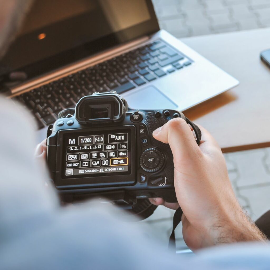 Close-up of person's hands holding a DSLR camera and scrolling through the settings, with a laptop in the background.