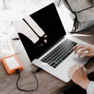 Macbook laptop, zoomed in to show typing hands and an orange, external hard drive plugged in