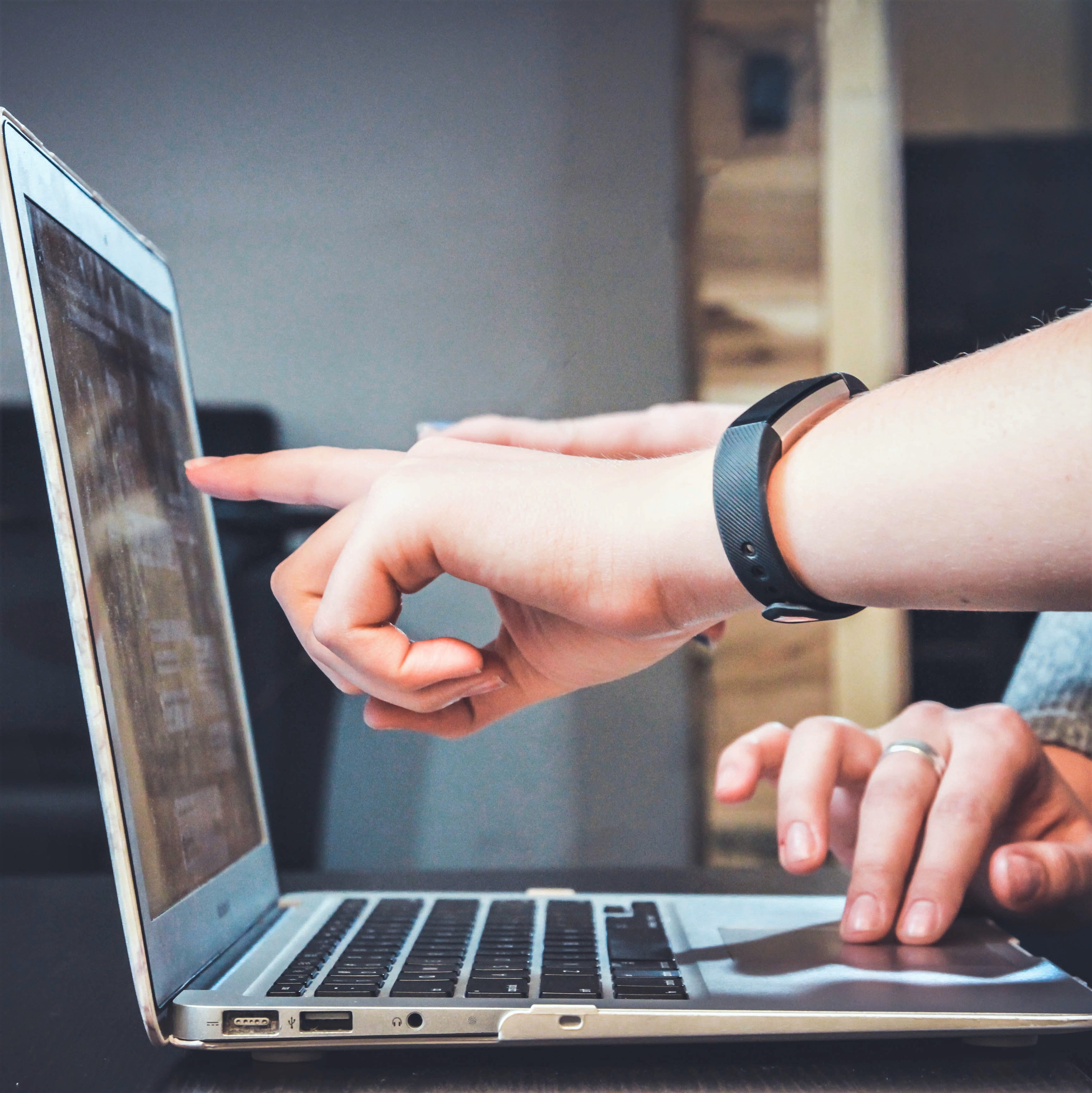 Open laptop with one hand pointing to something on the screen, and another person's hand scrolling on the trackpad. People's faces outside of view.