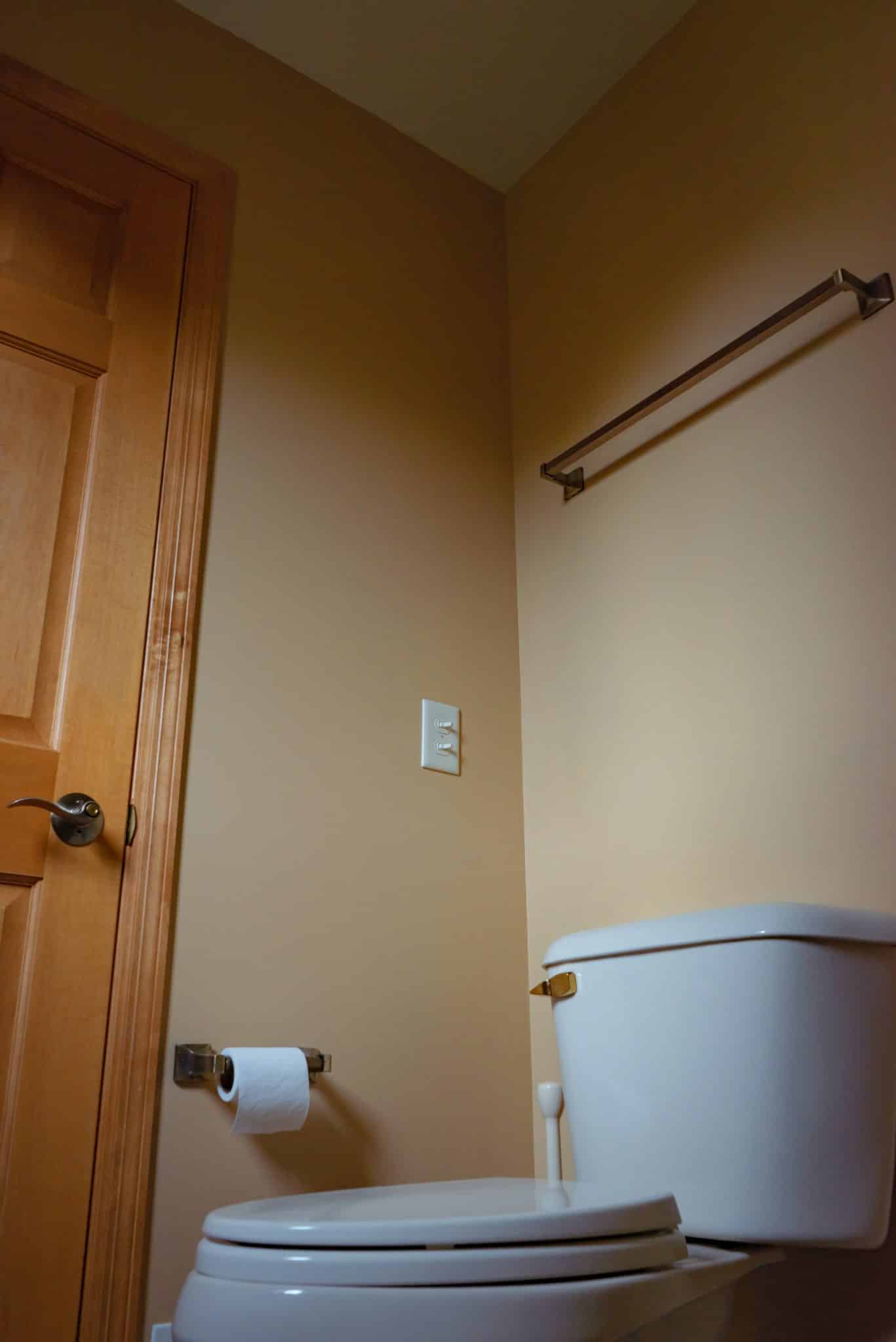 Photo of white toilet in bathroom with orange-brown walls and an oak-brown door. Camera angled from beneath toilet bowl-up, so ceiling and towel rack are visible.