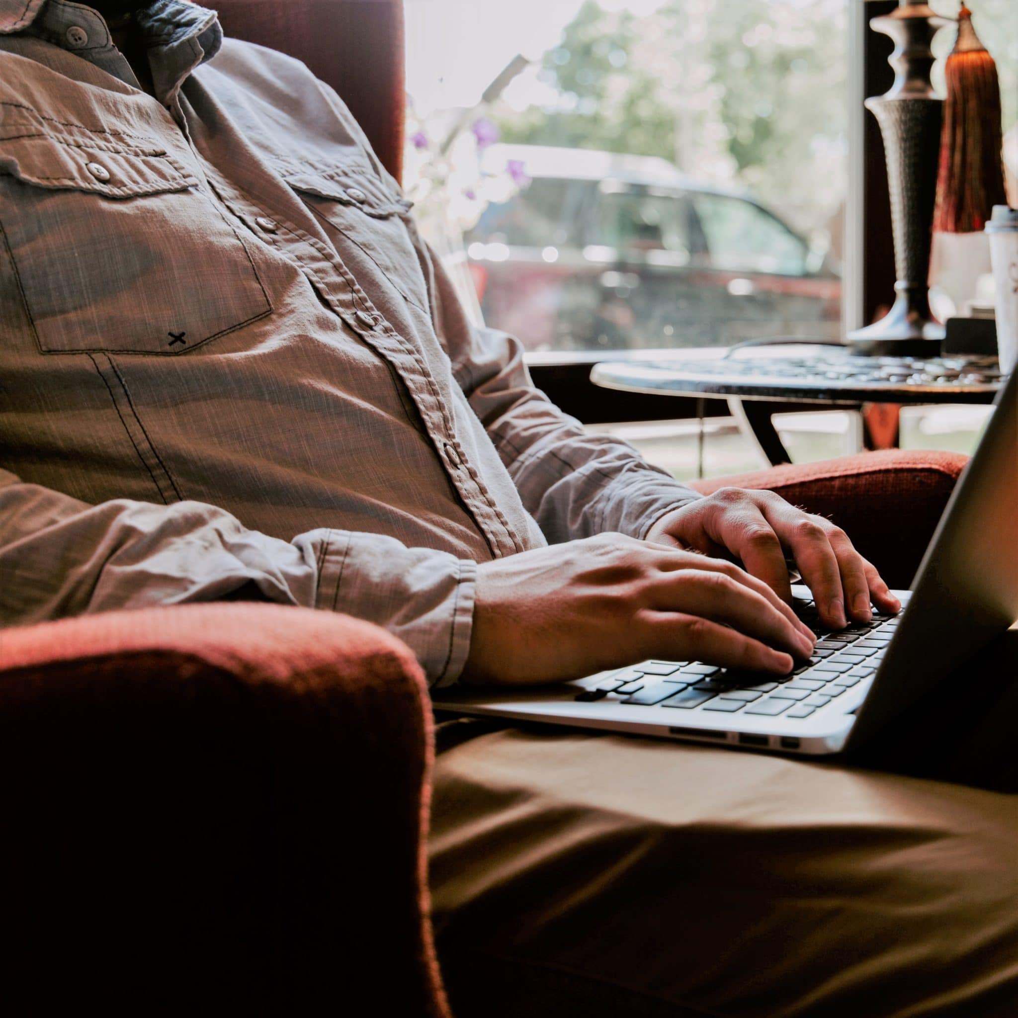 Man in button-down shirt sitting in red armchair, typing on laptop. Face not visible.