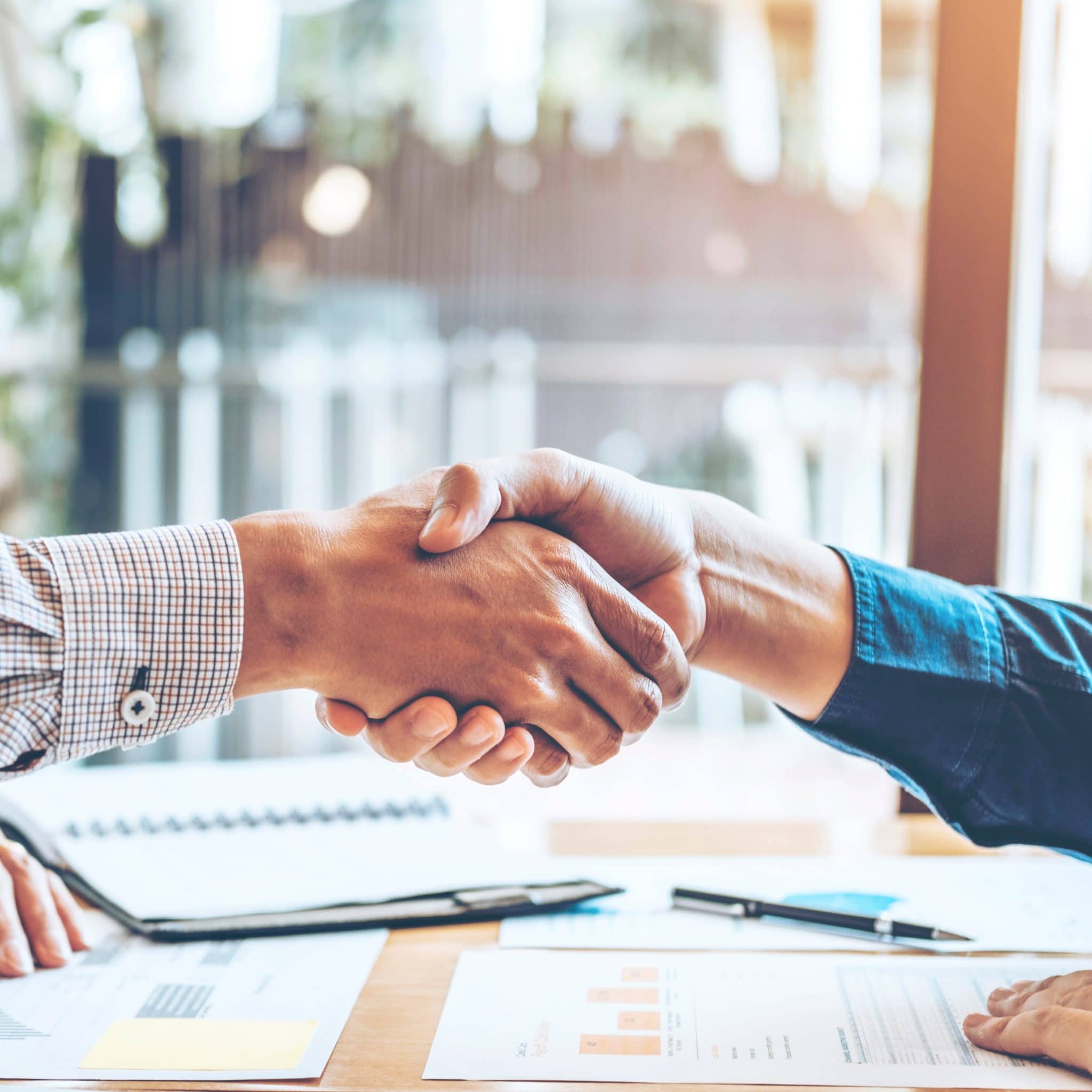 Photo of two men shaking hands at a table covered with paperwork. Zoomed in to show handshake and sleeved forearms.