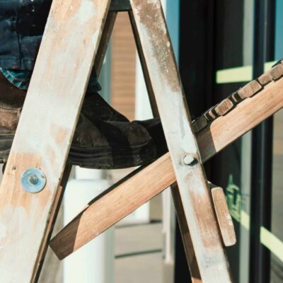 Zoomed in photo of man standing precariously on old, faded, wooden ladder, showing his brown boots and pantlegs