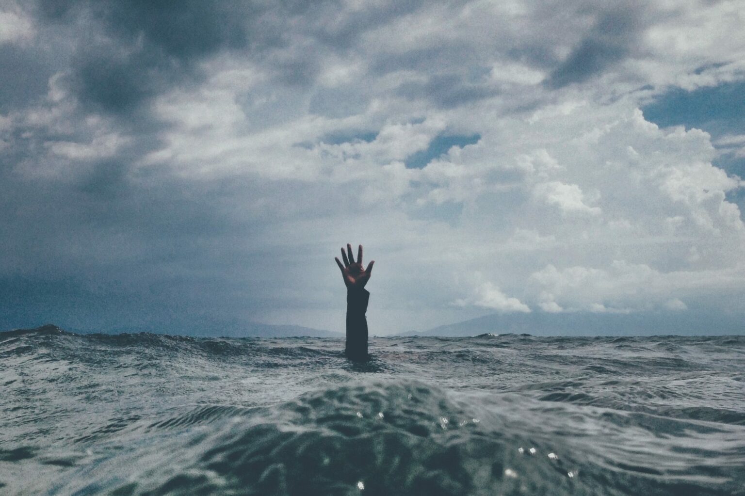nikko-macaspac-6SNbWyFwuhk-unsplash Rough, stormy, grey ocean with arm of drowning person reaching through surface for help.