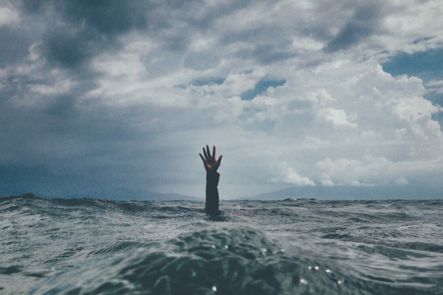 Rough, stormy, grey ocean with arm of drowning person reaching through surface for help.