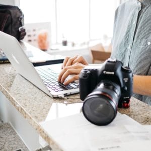 Person standing and typing on laptop with Canon camera to their left