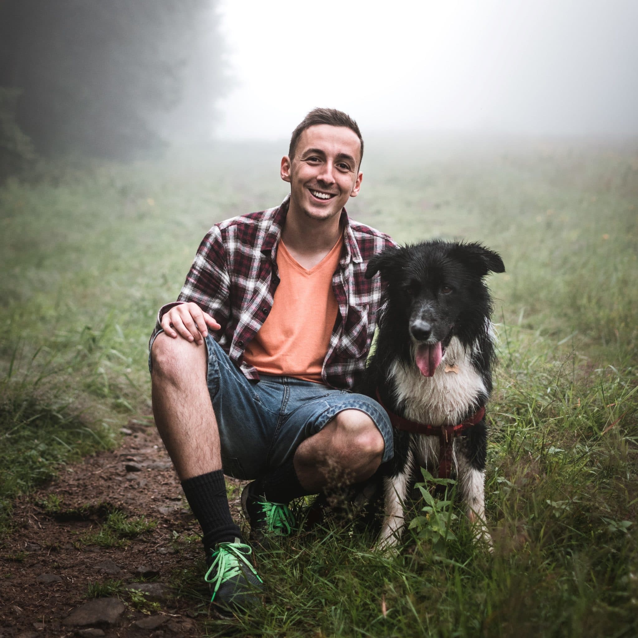 Casually dressed man standing on foggy trail with black and white dog by his side. Man is smiling, implying he and his dog are going for a walk.