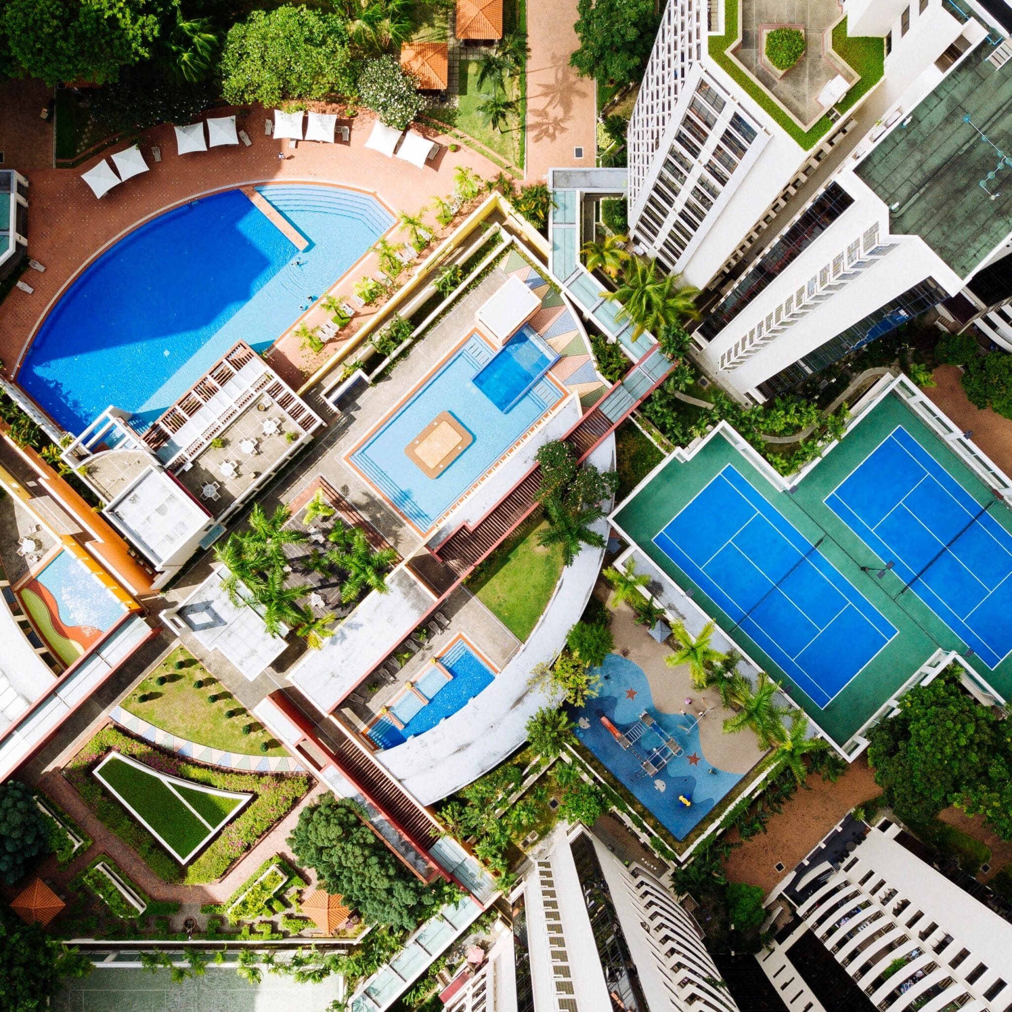 Birds-eye view of common spaces in an apartment or condo complex, featuring multiple pools, tennis courts, and outdoor lounging areas. The complex is set in a city with tall, modern buildings.