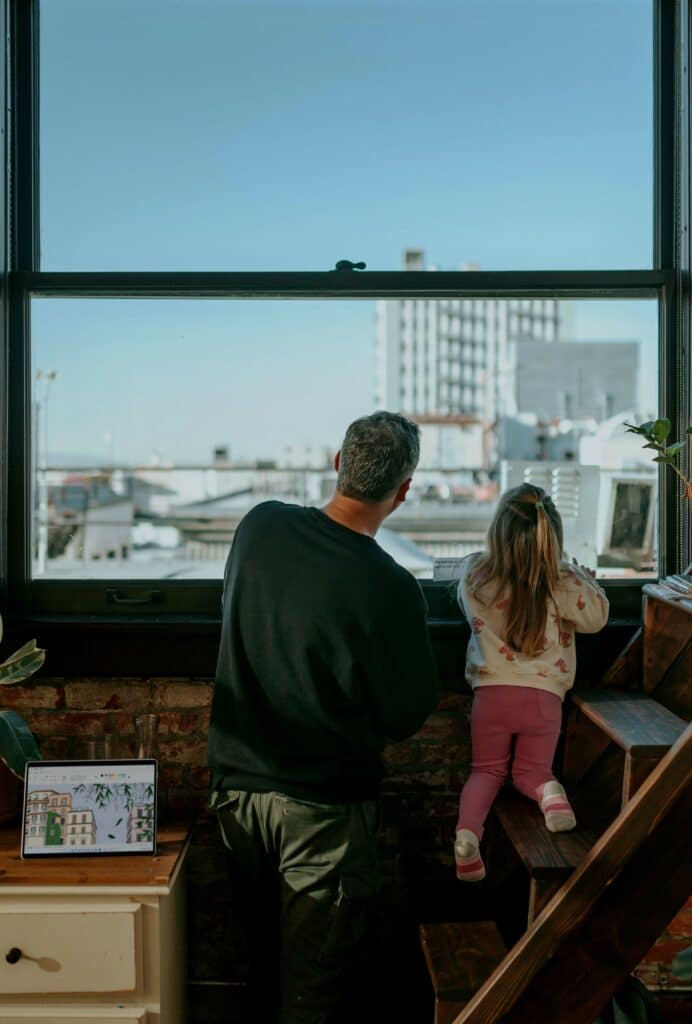 Man and young girl, about two years old, presumably his daughter, looking out a tall window. They appear to be in an apartment or condo, and they have an expansive view of the city with a blue, cloudless sky.