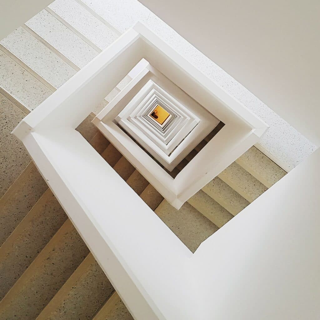 Camera panned downward of an indoor staircase. The staircase has white walls, tan steps, and descends in a square-shaped spiral for several flights. The image suggests an indoor staircase and common area for apartments and condos.