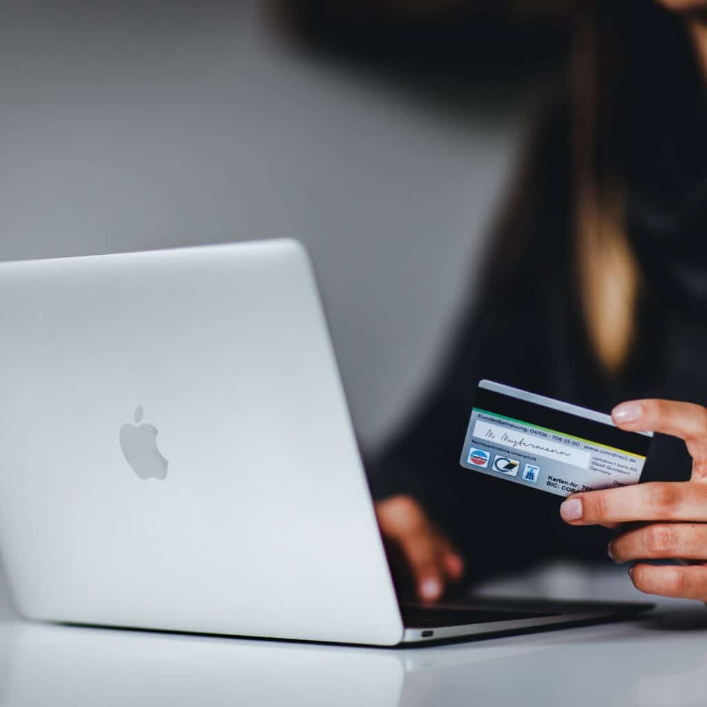 MacBook laptop on a white table, with person holding a credit card and typing on the laptop. Person's black shirt is blurry in the background, but face is not shown.