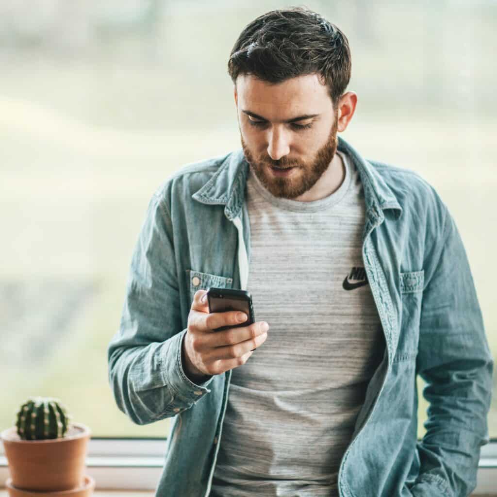 Man with short, brown hair and beard wearing blue, collared, long-sleeved shirt, looking down at phone in his hand