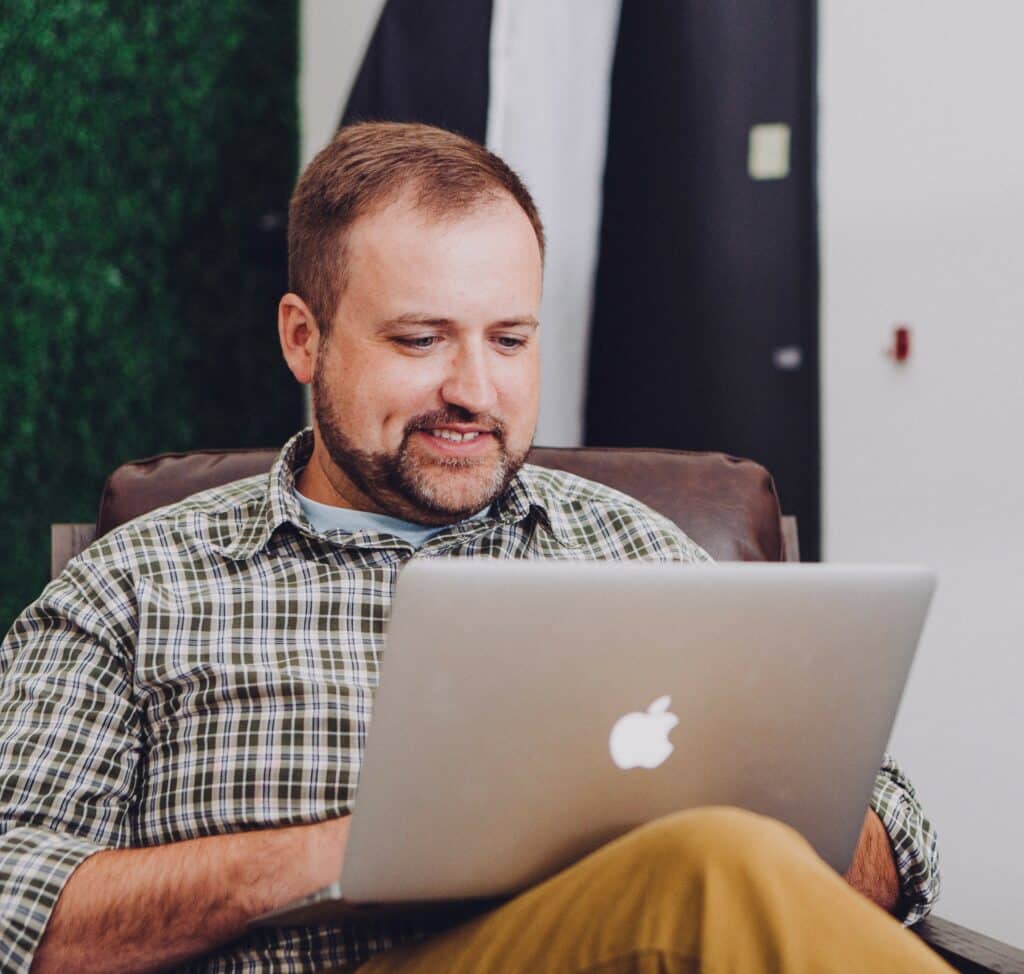 Man with brown hair and beard reclines in a brown, leather chair while smiling down at laptop in his lap, presumably something on the screen.