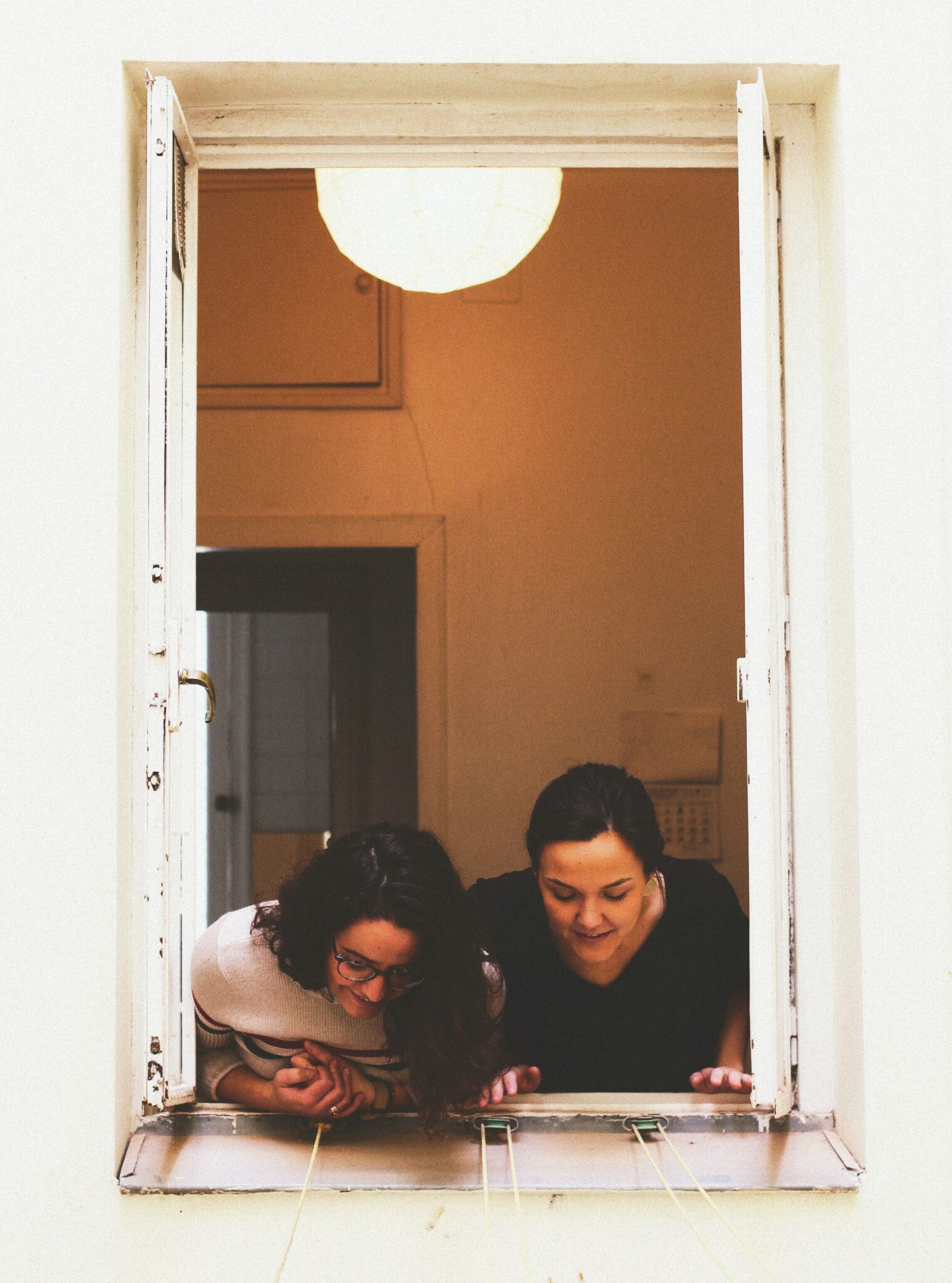 Faded, vintage-looking image of two young women with brown hair leaning out an open, white window in a building with white walls. The women are smiling and looking at something beneath the window but out of camera shot.