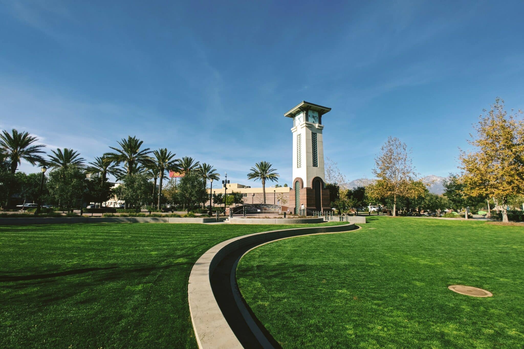 White, tall clock building on a green grass against a light blue sky, with palm trees in the background
