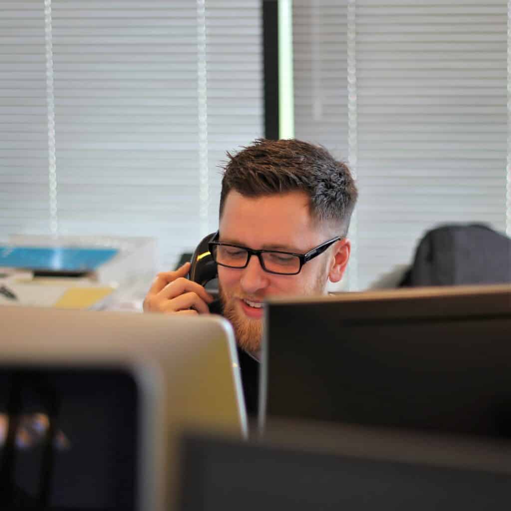 Man with short hair and glasses smiles while looking at dual computer monitors and talking into phone, perhaps providing support for an inspection report writing software. 