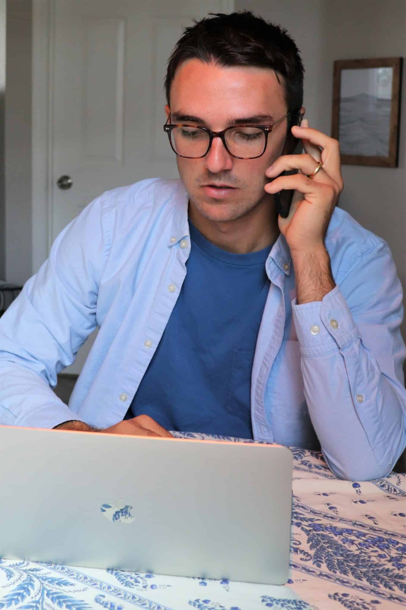 Man with short, dark hair sits at kitchen table typing on laptop, holding a phone to his ear with focused expression on his face