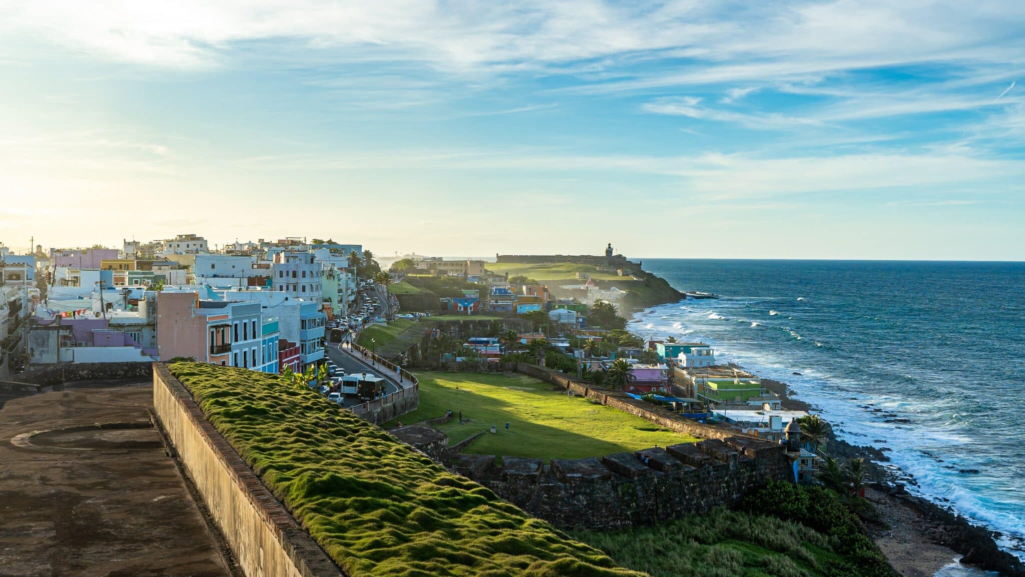 View of colorful buildings, dirt and asphalt walkways, lush, green grass, and the ocean off a cliffisde in San Juan, Puerto Rico