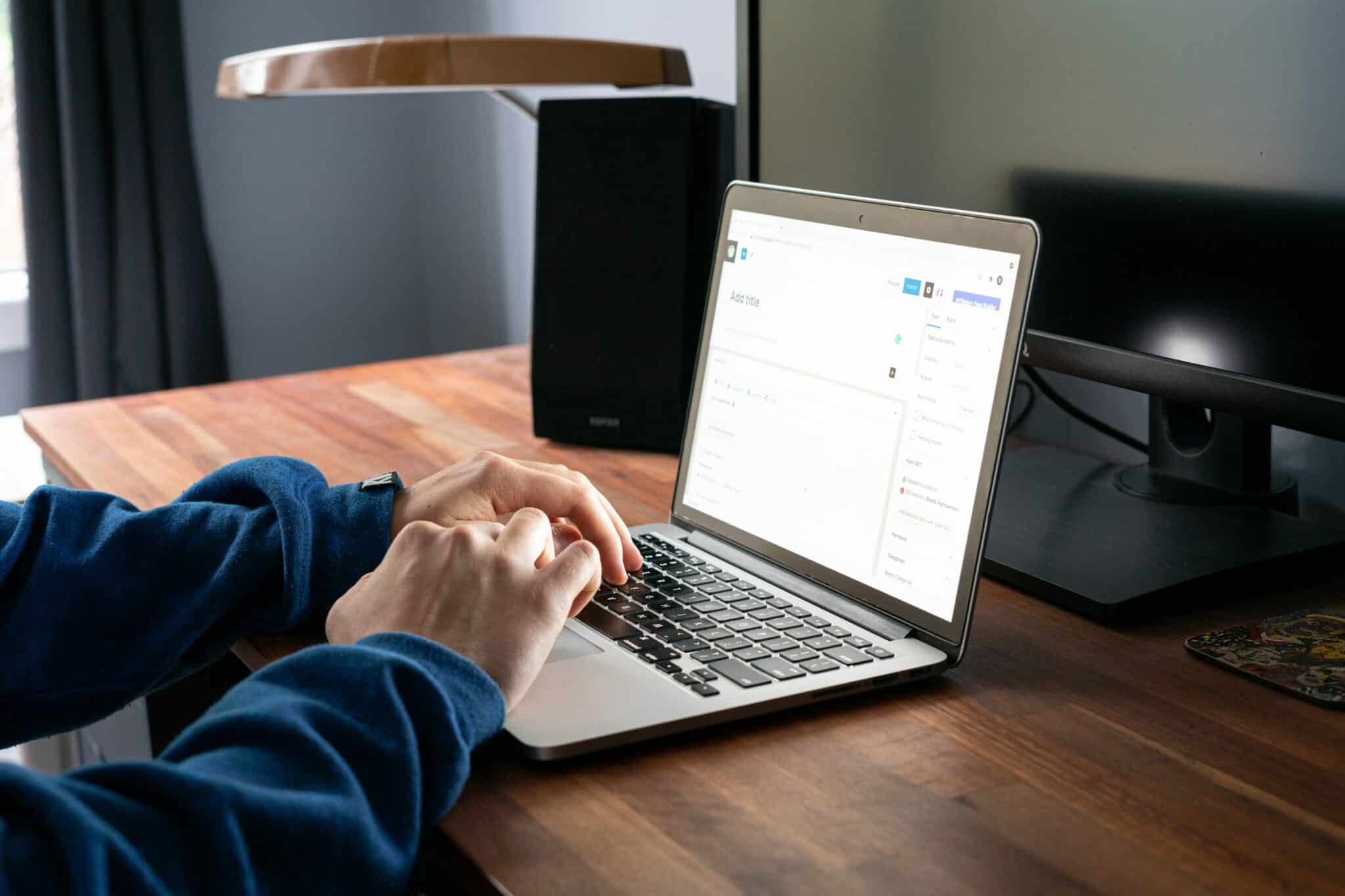 Zoomed in shot of person wearing a dark blue sweater, hands typing on a MacBook laptop at a wooden desk