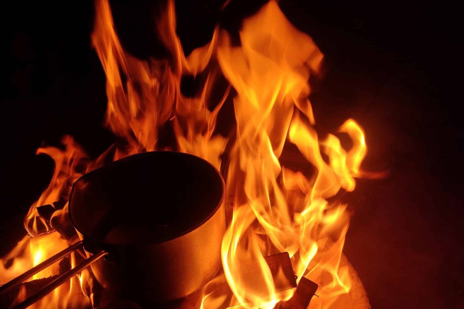 Metal cooking pot placed in a large, bright fire at nighttime