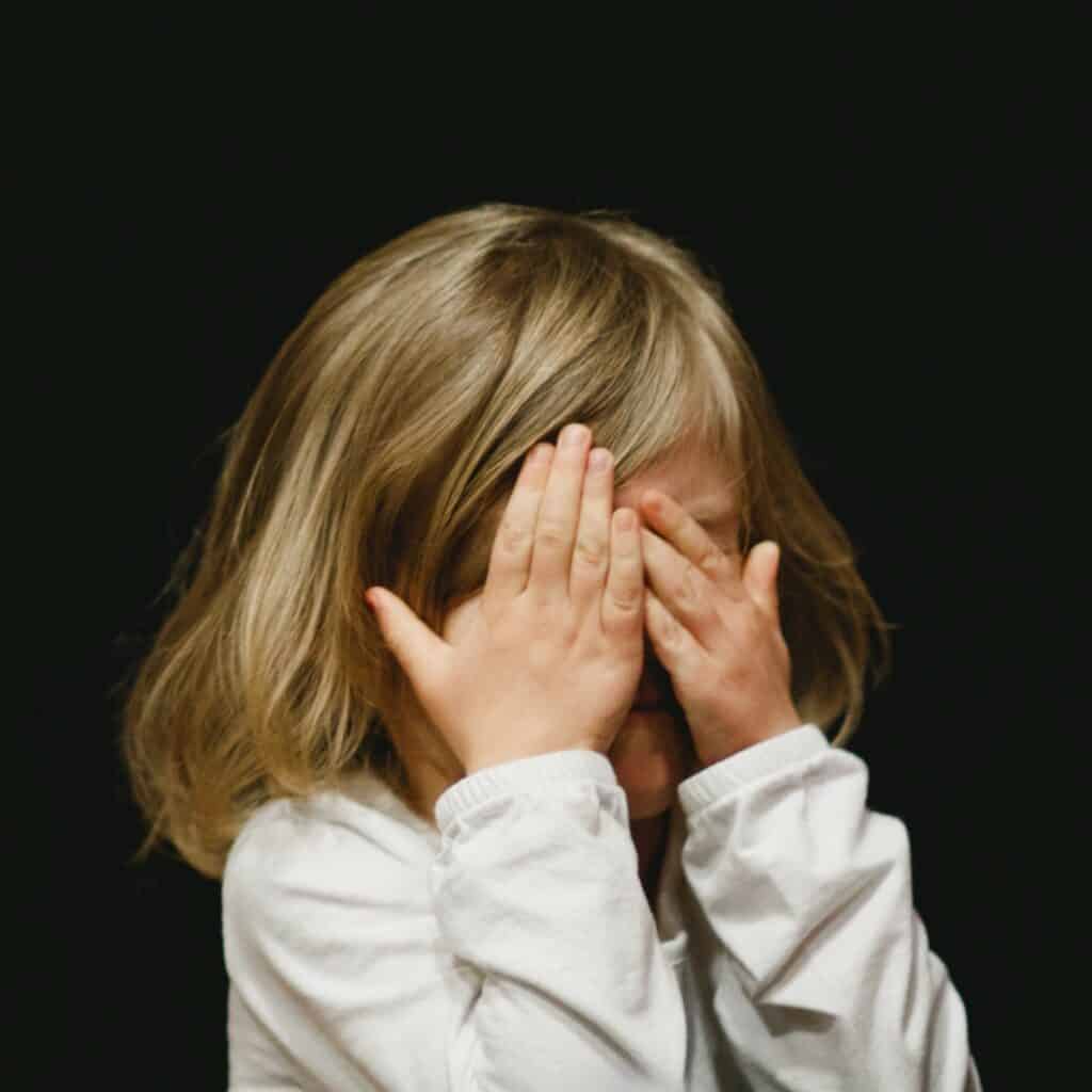 Young girl child with short blonde hair standing in front of a black background. Wearing long-sleeved, white shirt, covering face with hands as if about to cry. Represents admitting fault, guilt, or home inspector liability, as one should not do.