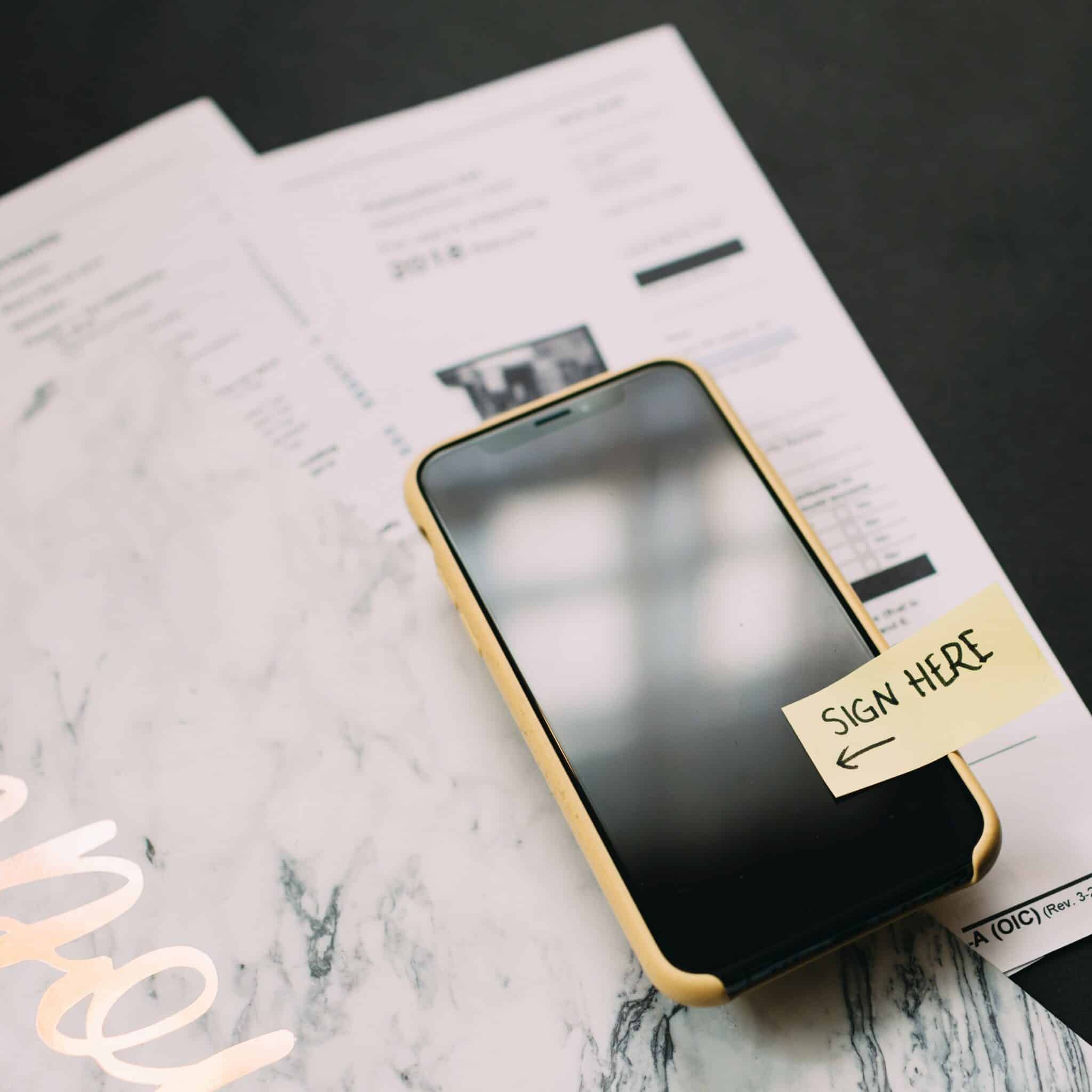 Smartphone with yellow case on top of paperwork and a marble-patterned folder on a black desk. Yellow sticky note on the phone screen says "Sign Here" with arrow pointing to the screen.