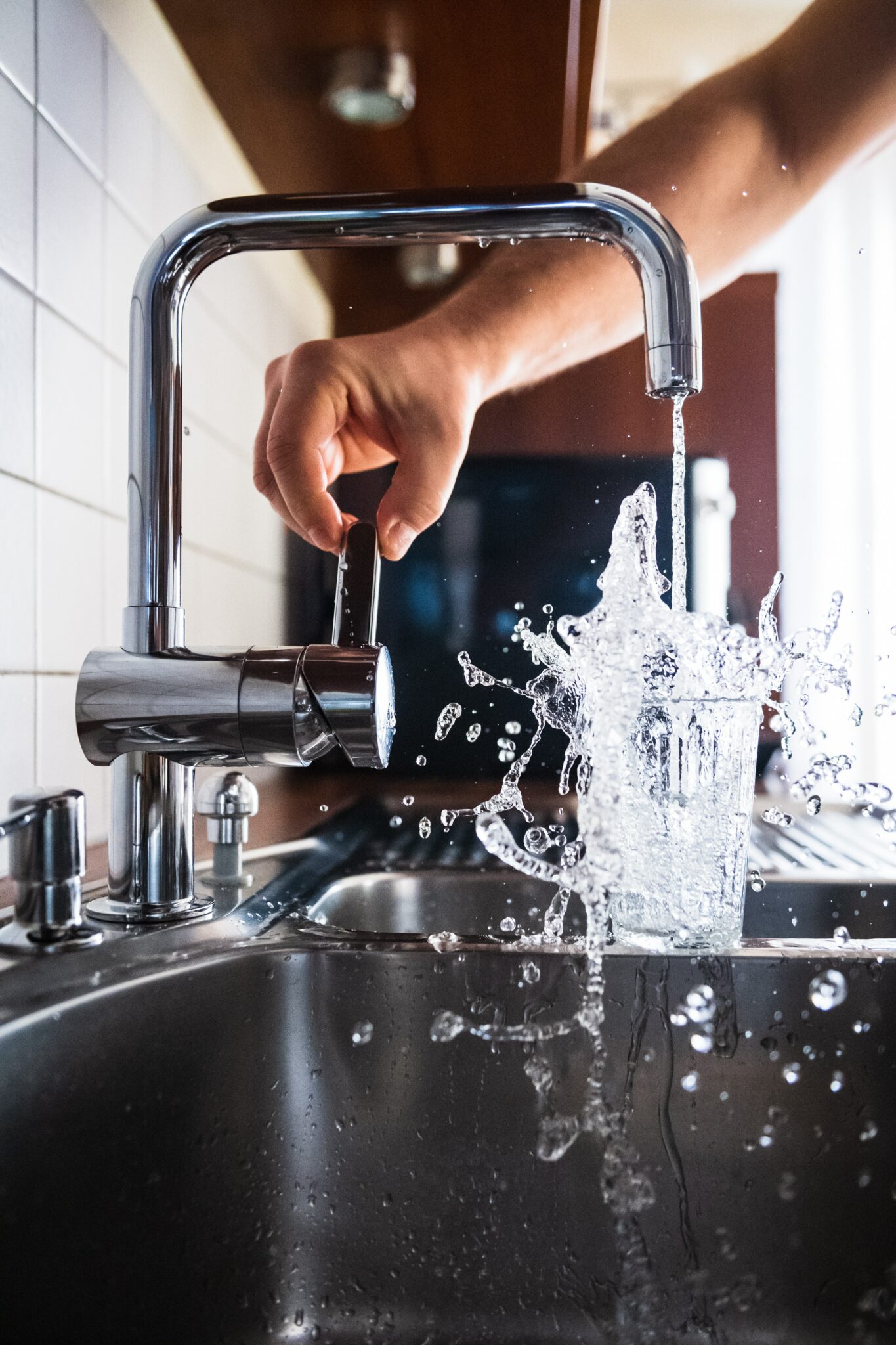 Close-up of person's hand turning on metal kitchen sink faucet and filling a glass cup with water. Water overflows and splashes into the sink.