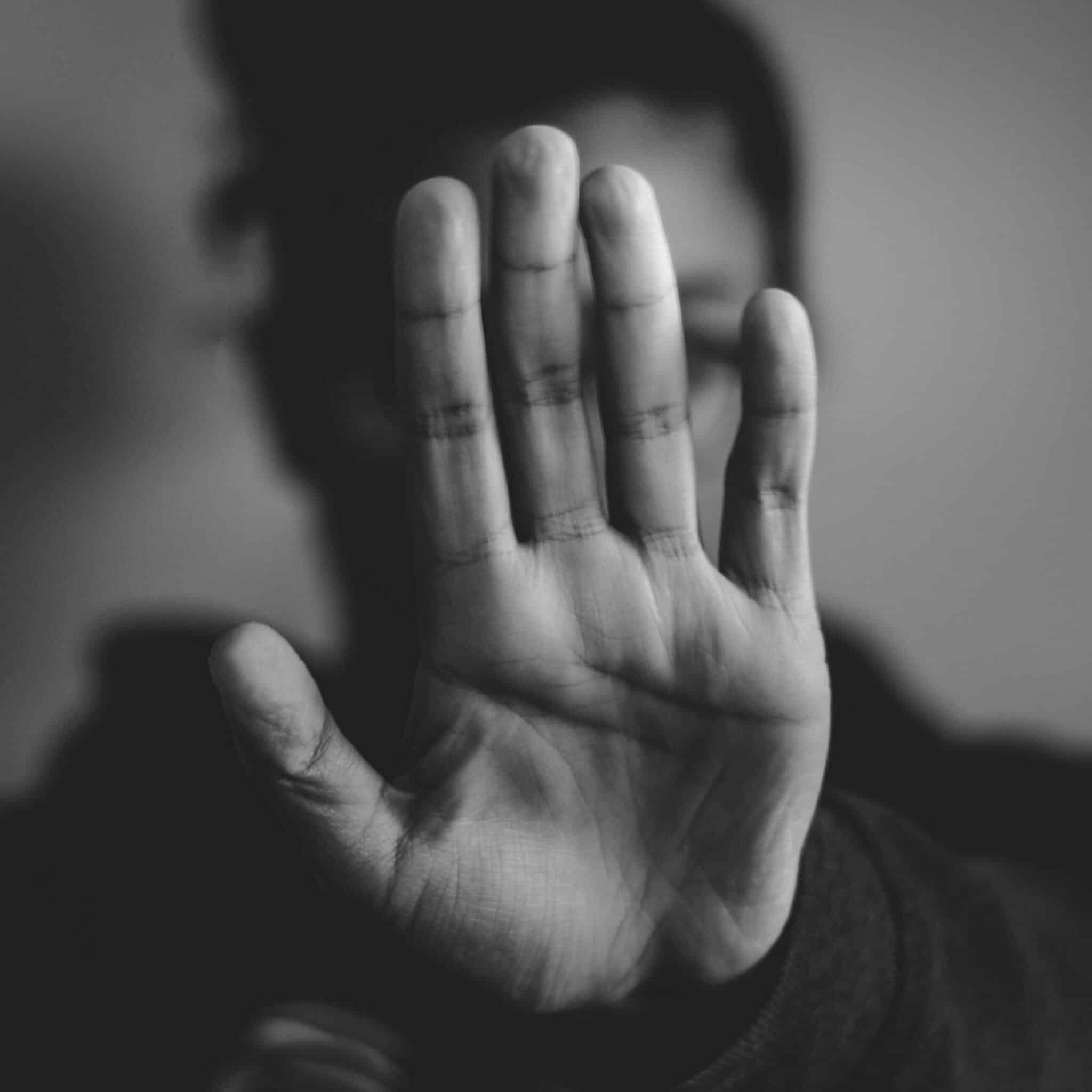 Low lighting image of person holding up hand to camera, as if saying "Stop." Person has dark clothes and hair and face is blurred in the background.