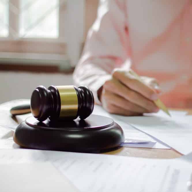 Zoomed in view of person's hand while writing on paper at a desk, with the person wearing a light-pink sleeved shirt and a legal gavel closer to camera on the desk