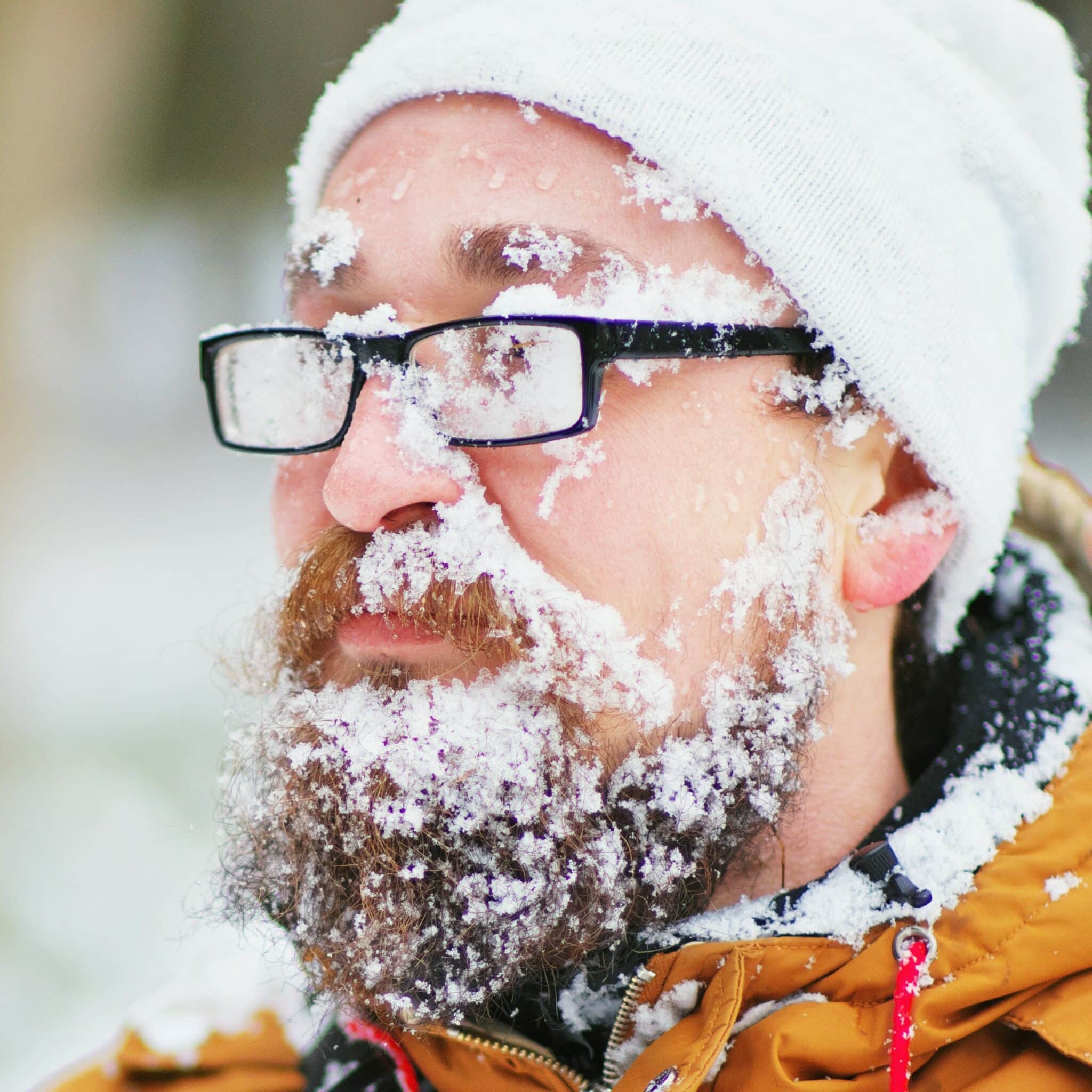 Middle-aged man wearing white beanie, black rectangular glasses, and a yellow snow coat, standing outside. He has light skin and a red beard. His beard and glasses are stuffed with snow, creating a comical appearance. It reminds inspectors to dress appropriately when dealing with home inspections during extreme weather.