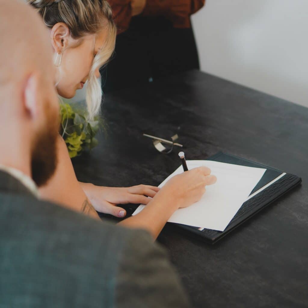 Blonde woman signs a blank piece of paper on a dark table, while a man in a business suit watches, blurred in the foreground.