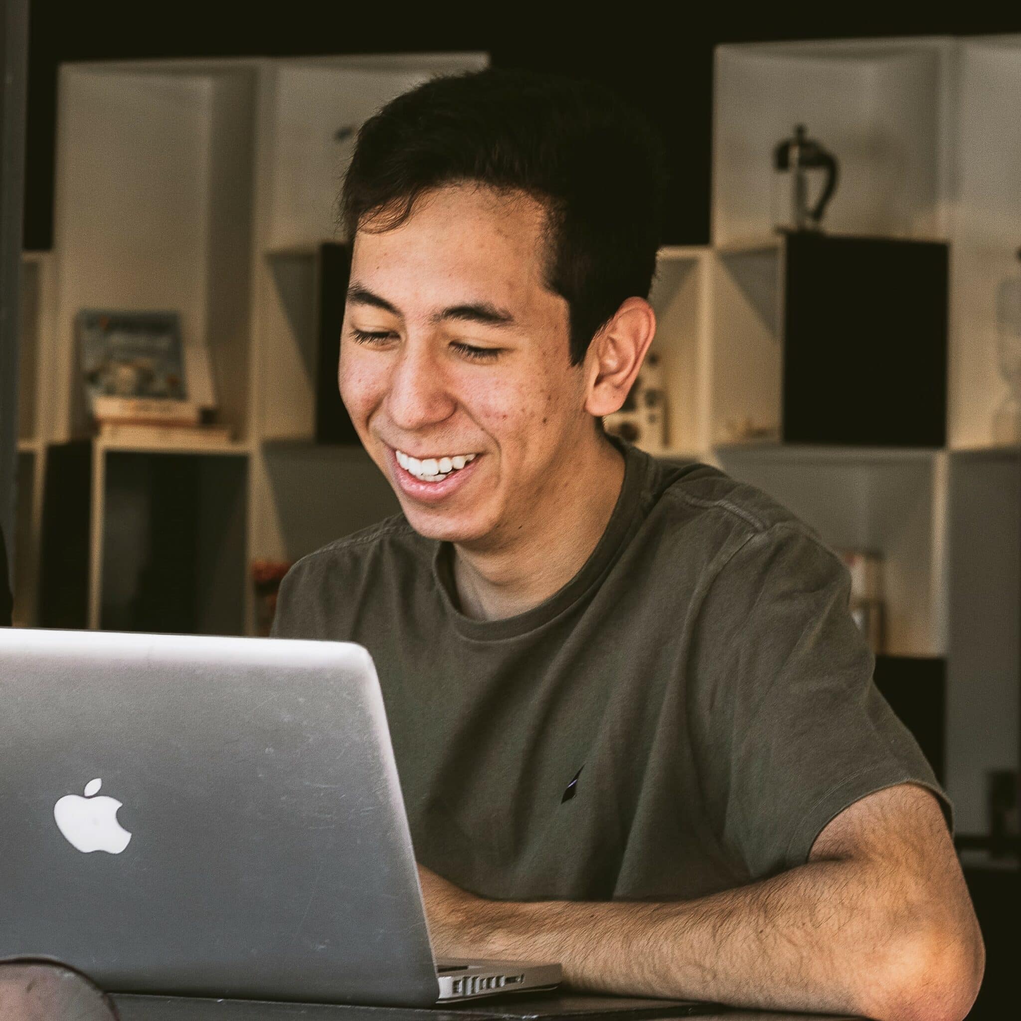 Young man with dark hair, wearing a t-shirt, smiling down at his Macbook laptop.