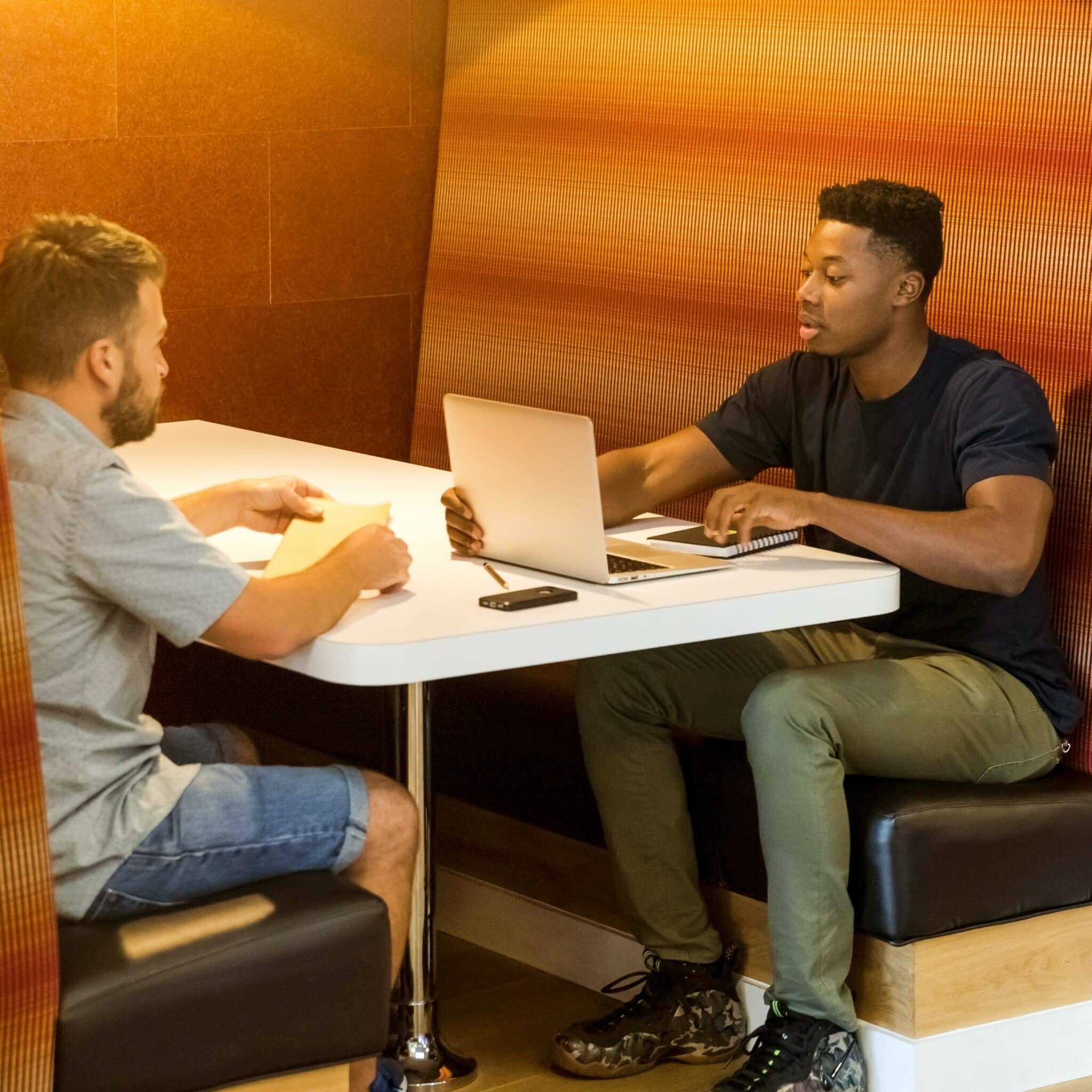 Older Caucasian man in t-shirt and shorts sits at booth, across from young Black man in green pants and a black, short-sleeved t-shirt, who's typing on a laptop. They appear to be discussing what's on his laptop, like a home inspection report or liability disclaimer.