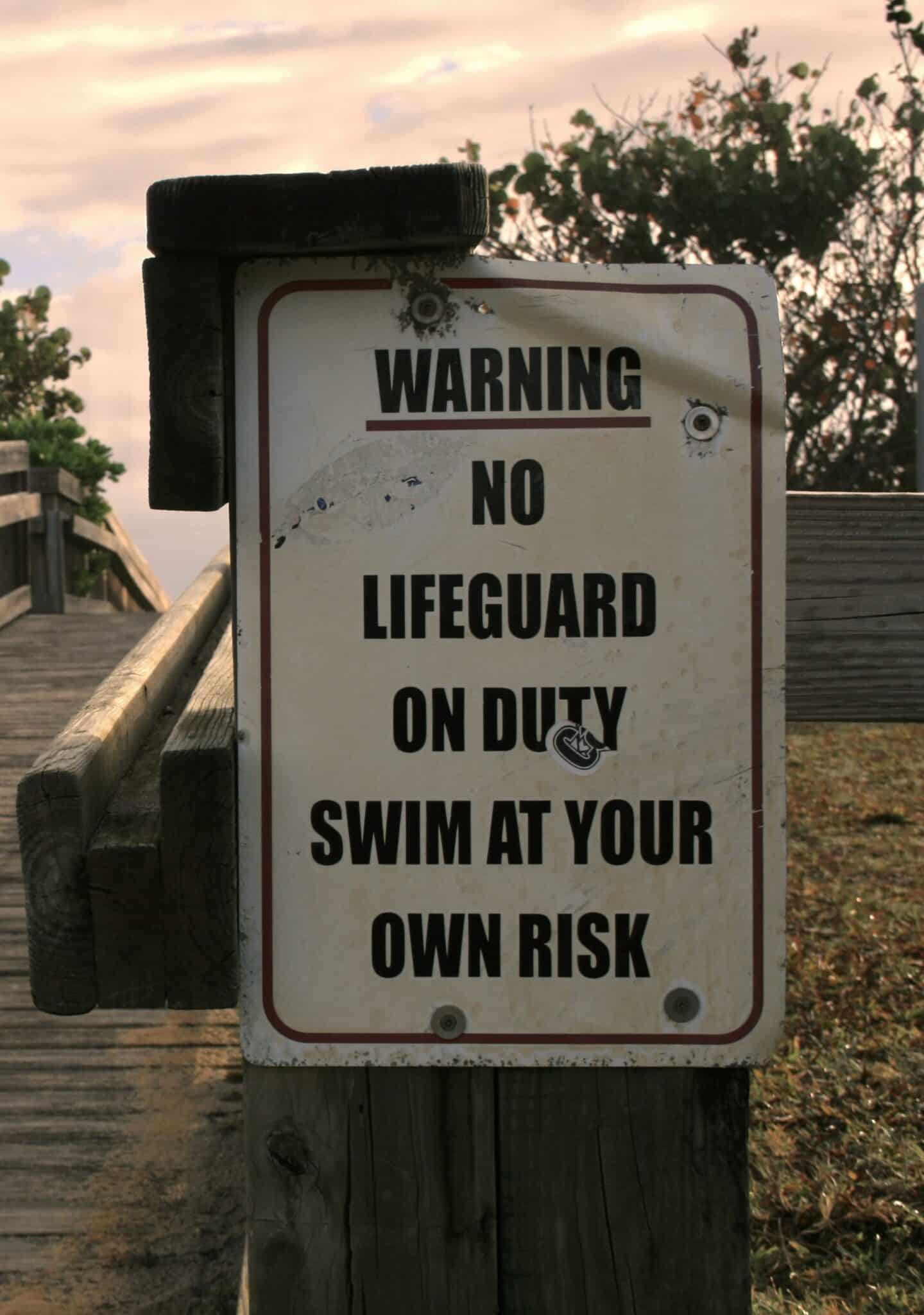 Liability disclaimer written on a white sign by a bridge, which says, "Warning-No lifeguard on duty. Swim at your own risk."