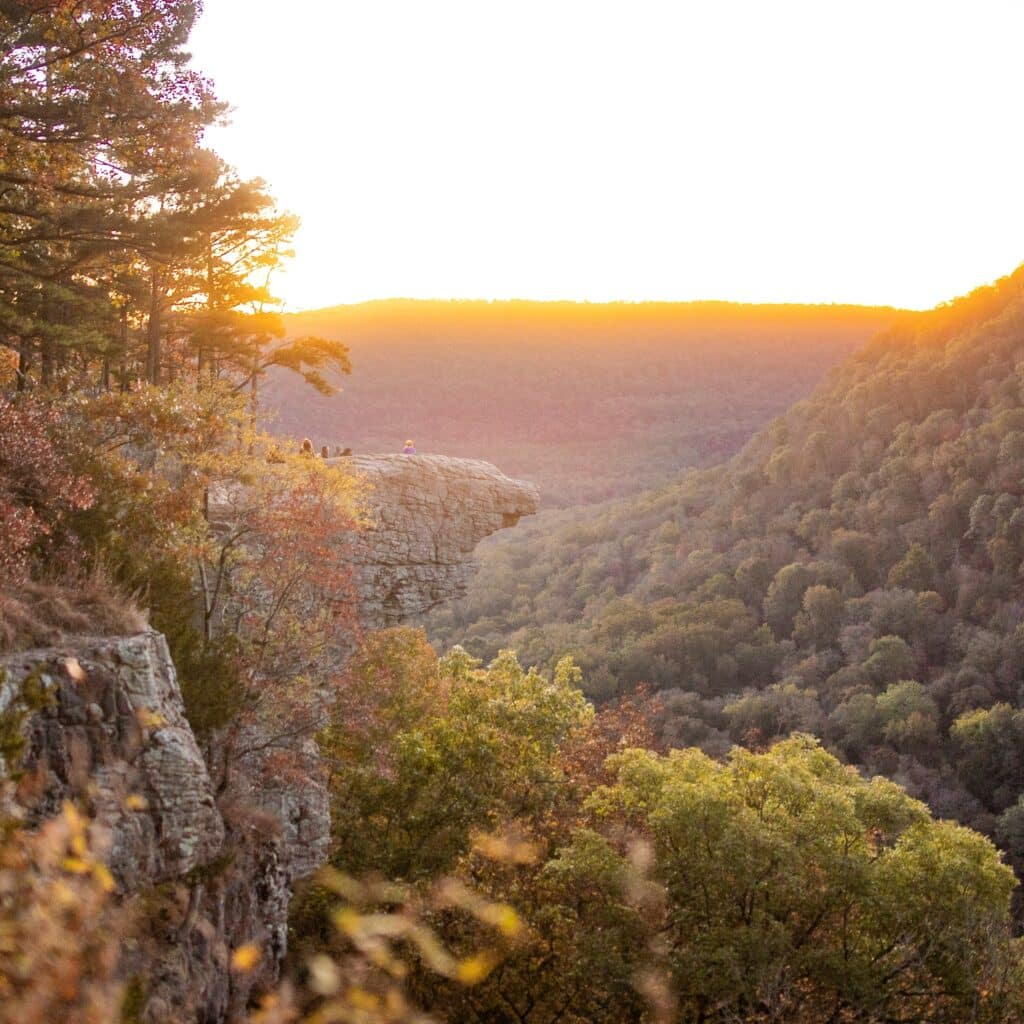 Sunset or sunrise at Whitaker Point, Hawksbill Crag, with fall foliage and a cliffside rock jutting out to a valley below. Hikers sit on the rock looking away from the camera.