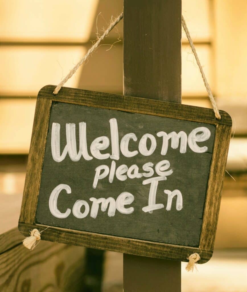 Green, small chalkboard sign hanging by a string from a post. Writing says "Welcome Please Come In," signifying a realtor and home inspector agreeing to do a walkthrough together.