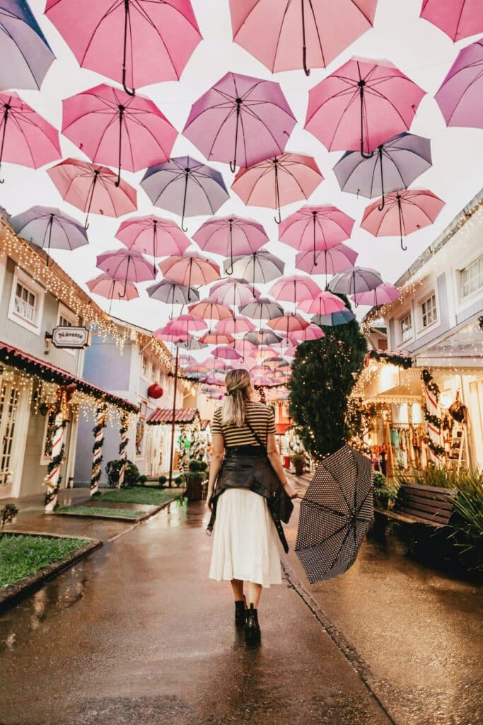 Woman holding umbrella and walking under dozens of pink and grey umbrellas in the sky. She walks through a narrow street with Christmas decorations on both sides.