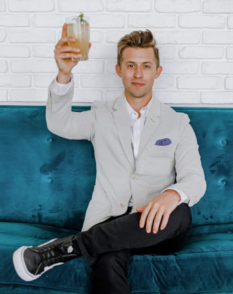 Man in business suit and sneakers sitting on a blue couch, looking at camera, and holding up mixed cocktail drink. Illustrates realtor and home inspector conflicts with agents wanting to attend the inspection.