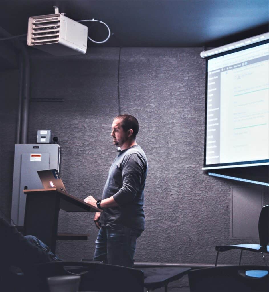 Realtor and home inspector presentation, featuring a man in a button-up shirt and jeans presenting before a crowd with a projector behind him.