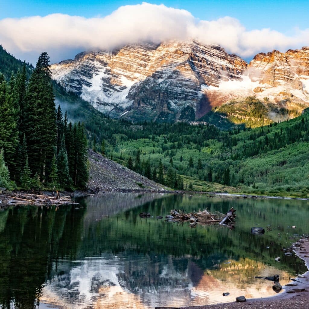 Lush, green pine trees, a shallow but wide river, and snowy mountains of Maroon Bells, Colorado, looking up at a blue sky with clouds.