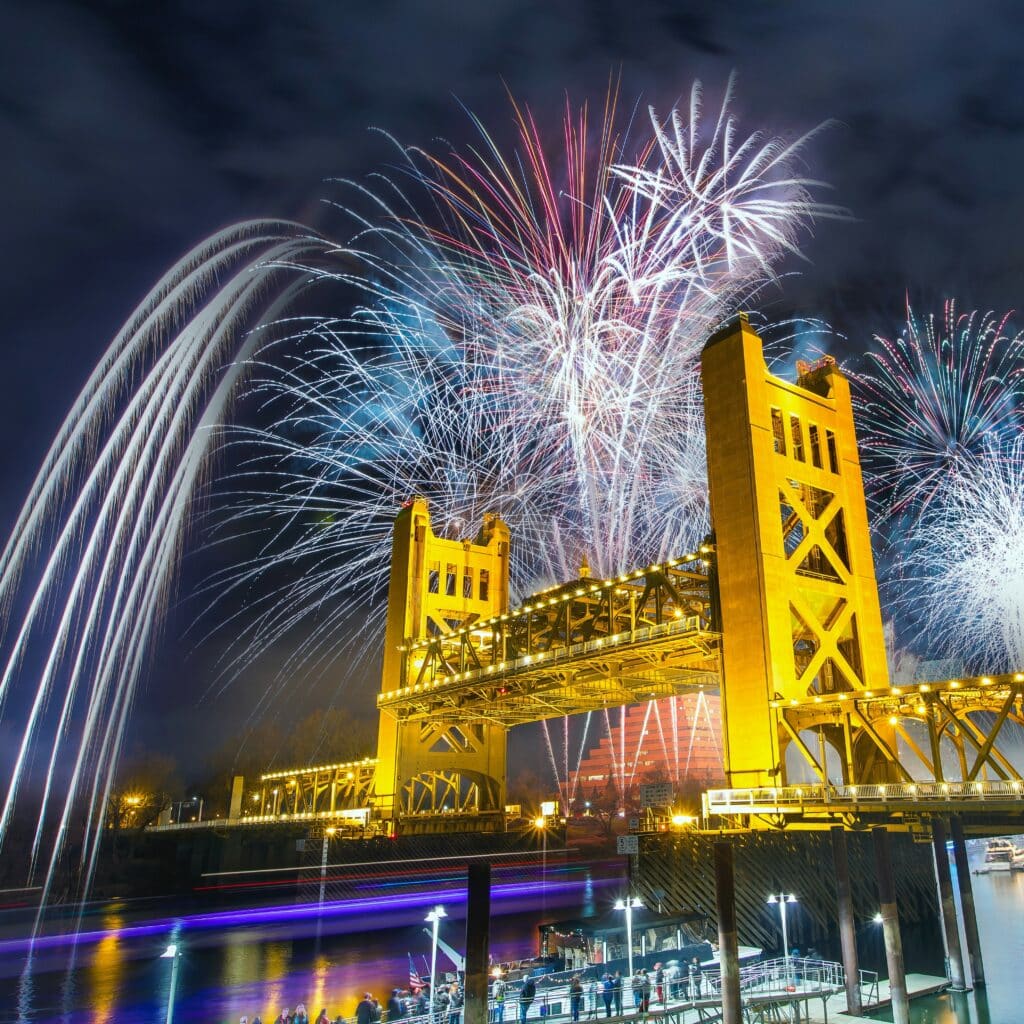 New Year’s Eve fireworks show at Sacramento’s Tower Bridge at night time, a crowd watching by the Sacramento river.