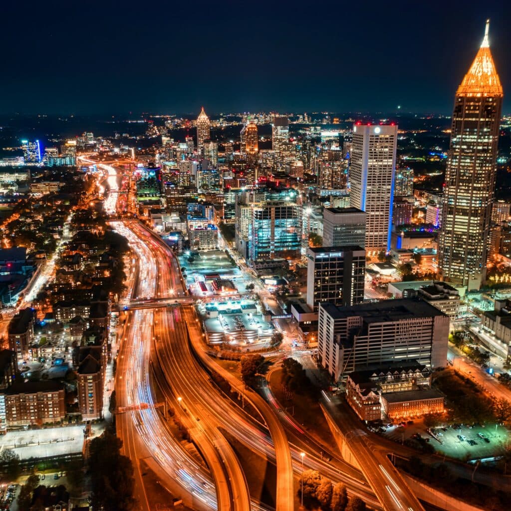Cityscape of Atlanta, GA at night, featuring an indigo sky, a tall, pointed skyscraper, and long freeways.