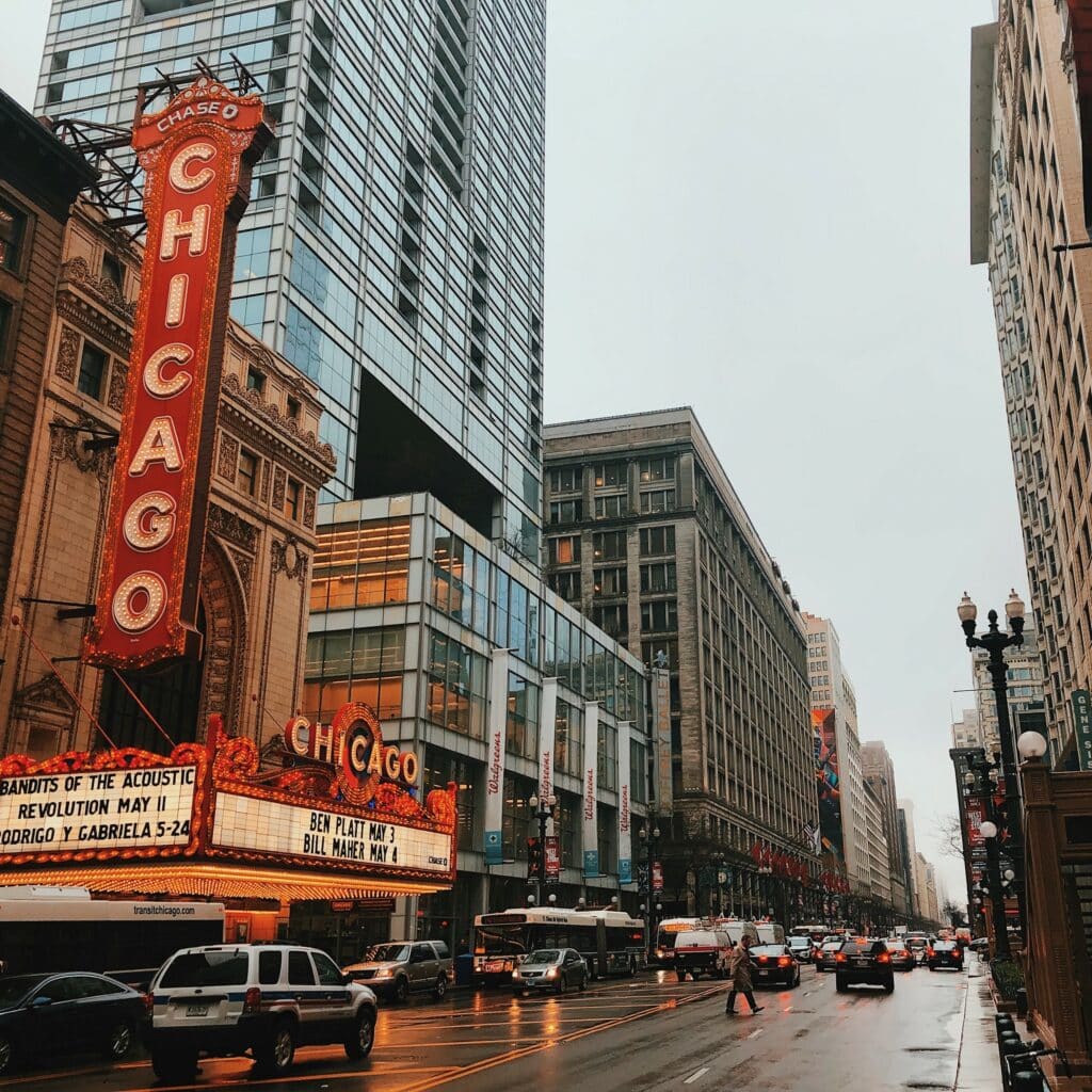 Urban scene in Chicago, Illinois, featuring a theater with a tall “Chicago” sign hanging vertically and several cars driving through a busy, city street on an overcast day.