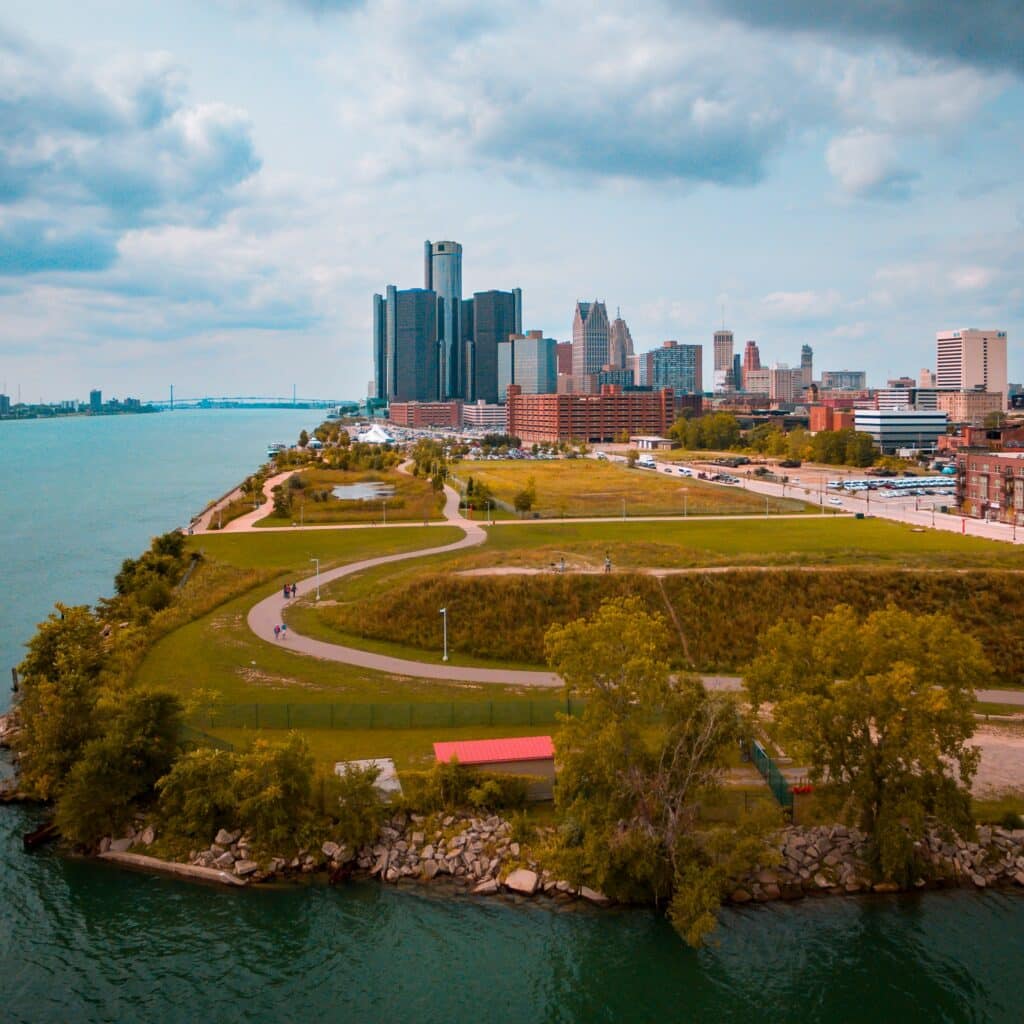 Grassy park running along the Detroit River in Michigan, with city skyscrapers and other buildings in the background.