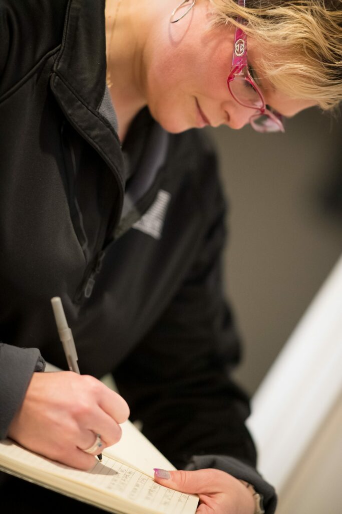 Woman wearing glasses and black polo writes on a notepad with pen to follow along before-you-go practices for home inspectors.