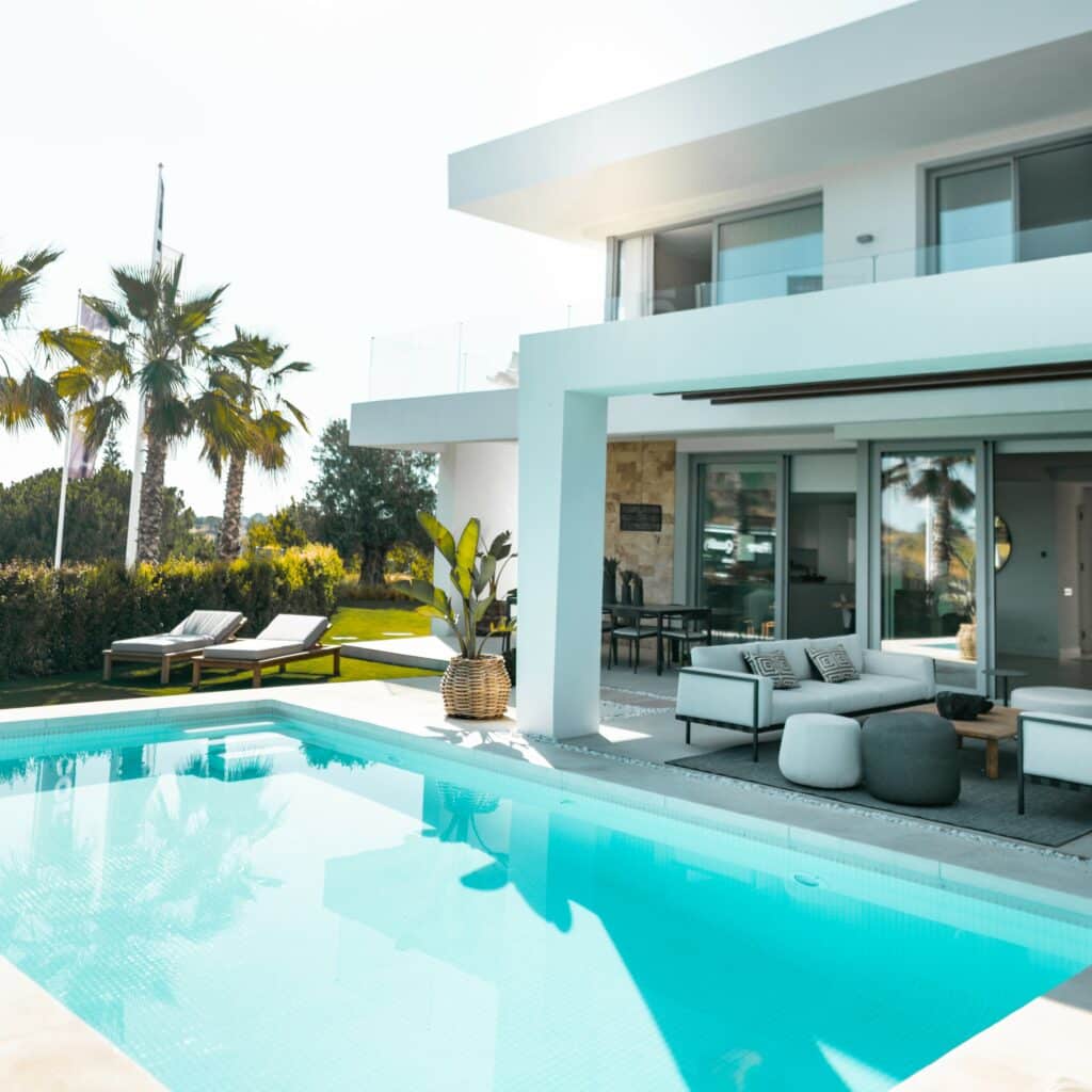 Modern backyard pool during daytime with palm trees and lawn furniture surrounding.