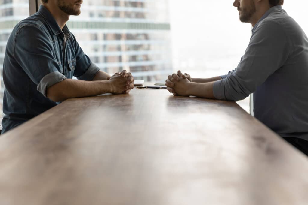 Two men, a realtor and home inspector, sitting across from each other at a long table. One man has a casual shirt with sleeves rolled up, the other has a more formal, button up shirt.