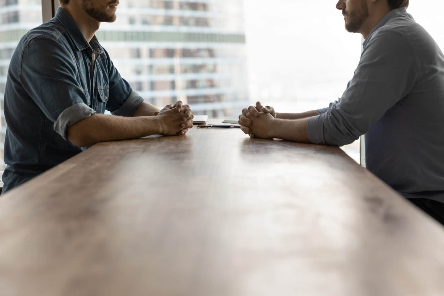 Determined,Young,Businessmen,Sit,Opposite,At,Desk,Face,Each,Other Two men, a realtor and home inspector, sitting across from each other at a long table. One man has a casual shirt with sleeves rolled up, the other has a more formal, button up shirt.