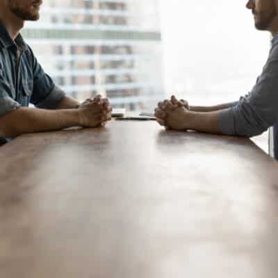 Two men, a realtor and home inspector, sitting across from each other at a long table. One man has a casual shirt with sleeves rolled up, the other has a more formal, button up shirt.