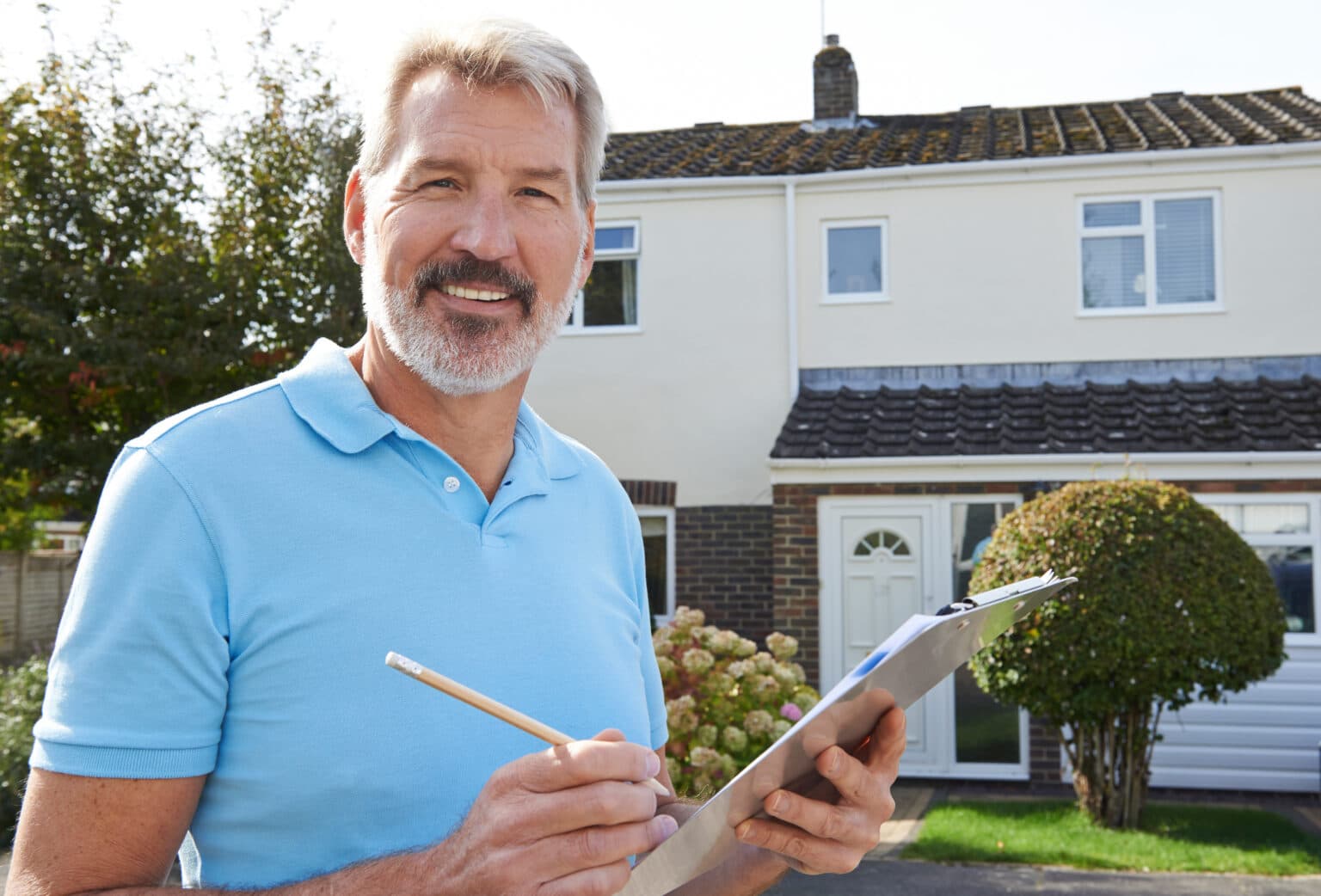 Man with grey hair and beard smiles at the camera while holding a clipboard in front of a home, presumably exhibiting the best course of action after a home inspection.